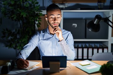 Sticker - Young hispanic man working at the office at night with hand on chin thinking about question, pensive expression. smiling and thoughtful face. doubt concept.