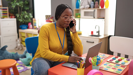 african woman preschool teacher using laptop speaking on the phone at kindergarten