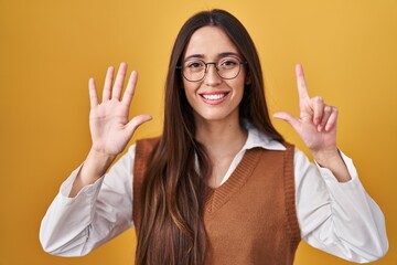 Sticker - Young brunette woman standing over yellow background wearing glasses showing and pointing up with fingers number seven while smiling confident and happy.