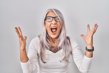 Poster - Middle age woman with grey hair standing over white background crazy and mad shouting and yelling with aggressive expression and arms raised. frustration concept.