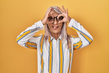 Poster - Middle age woman with grey hair standing over yellow background wearing glasses doing ok gesture like binoculars sticking tongue out, eyes looking through fingers. crazy expression.