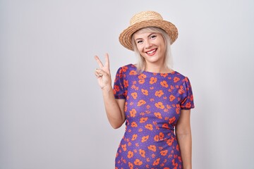 Poster - Young caucasian woman wearing flowers dress and summer hat showing and pointing up with fingers number two while smiling confident and happy.