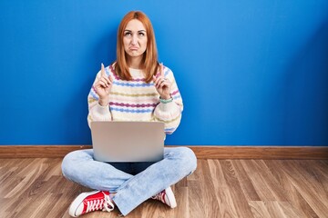 Wall Mural - Young woman using laptop at home sitting on the floor pointing up looking sad and upset, indicating direction with fingers, unhappy and depressed.