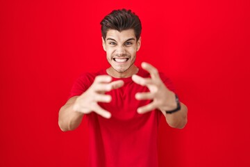 Wall Mural - Young hispanic man standing over red background shouting frustrated with rage, hands trying to strangle, yelling mad