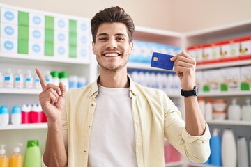 Sticker - Young hispanic man at pharmacy drugstore holding credit card smiling happy pointing with hand and finger to the side