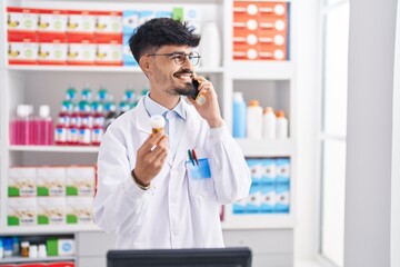 Wall Mural - Young hispanic man pharmacist talking on smartphone holding pills at pharmacy