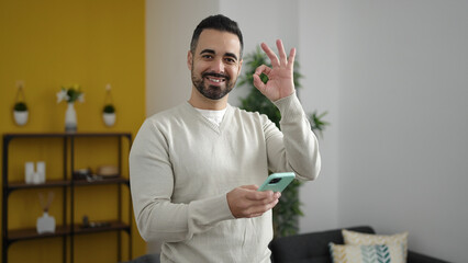 Canvas Print - Young hispanic man using smartphone doing ok gesture at home