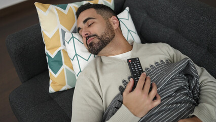 Canvas Print - Young hispanic man holding tv remote control lying on sofa sleeping at home