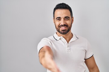 Sticker - Young hispanic man with beard wearing casual clothes over white background smiling friendly offering handshake as greeting and welcoming. successful business.