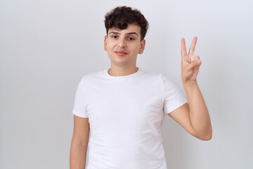 Canvas Print - Young non binary man wearing casual white t shirt showing and pointing up with fingers number two while smiling confident and happy.