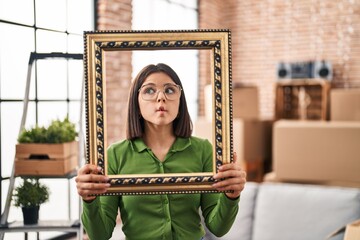 Canvas Print - Young hispanic woman at new home holding empty frame making fish face with mouth and squinting eyes, crazy and comical.