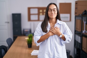 Canvas Print - Young hispanic woman at the office smiling with hands on chest with closed eyes and grateful gesture on face. health concept.