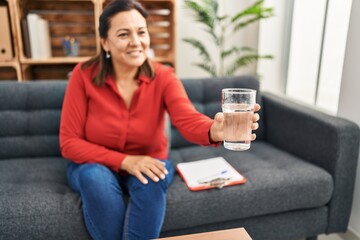 Wall Mural - Middle age hispanic woman psychologist offering glass of water at psychology center