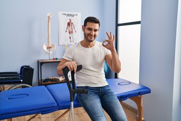 Sticker - Young hispanic man with beard wearing crutches at rehabilitation clinic doing ok sign with fingers, smiling friendly gesturing excellent symbol