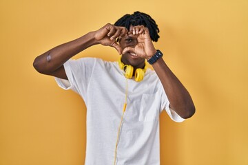 Sticker - Young african man with dreadlocks standing over yellow background doing heart shape with hand and fingers smiling looking through sign