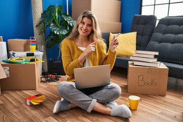 Canvas Print - Young woman sitting on the floor at new home using laptop smiling and looking at the camera pointing with two hands and fingers to the side.