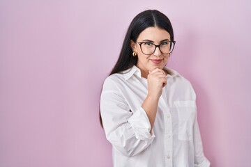 Sticker - Young brunette woman standing over pink background looking confident at the camera with smile with crossed arms and hand raised on chin. thinking positive.