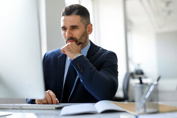 Concentrated middle aged male entrepreneur working with computer, looking at screen and thinking, office interior