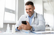 © Prostock-studio - Smiling male doctor in medical uniform sitting at desk in clinic, looking at phone screen, consulting client online