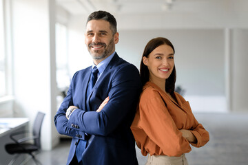 Two successful businesspeople standing back to back with arms crossed and smiling at camera, posing in modern office