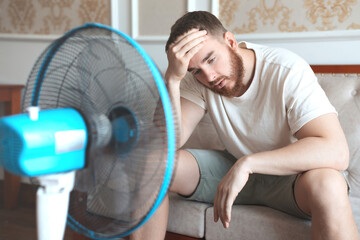 Young bearded man using electric fan at home, sitting on couch cooling off during hot weather, suffering from heat, high temperature