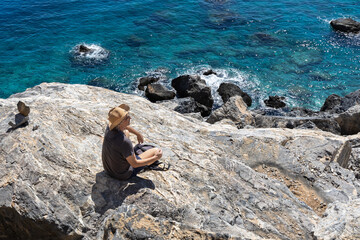 A man in a hat sits on a stone near the turquoise sea, Greece, Crete