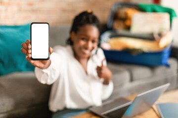 Wall Mural - Travel offer concept. Happy black lady showing cellphone with blank screen, sitting with suitcase at home, mockup