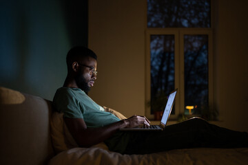 Focused African American programmer man in dark room lying on bed work on laptop. Workaholic freelancer black guy sitting comfortably with computer on laps working on graphic design for app at night