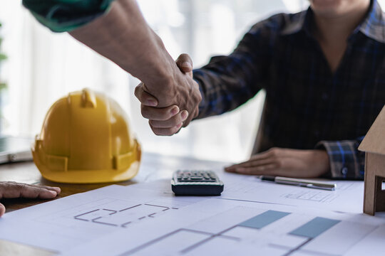 yellow hard hat on table and house design print design with construction team handshake greeting sta
