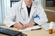 © DragonImages - Close-up of young male doctor in lab coat making notes in medical card of online patient or writing down prescriptions during consultation