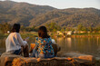 © aumnat - Two young woman relaxing with using mobile phone while sitting  beside lake in a nature park.