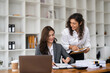 © Natee Meepian - Two diverse female smiling while working together at a boardroom table during a meeting in a modern office