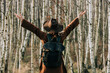 © Westend61 - Woman wearing backpack standing with arms raised looking at birch trees