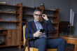 © artmim - Elegant elderly successful businessman in blue suit and glasses looking on his mobile phone, sitting in office
