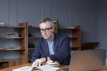 Portrait of mature businessman in glasses and blue suit sitting by laptop and pensivly reading the book in his office