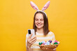 © sementsova321 - Portrait of smiling joyful woman wearing bunny ears holding easter eggs isolated over yellow background, using mobile phone, massaging with friends, congratulating with holiday.