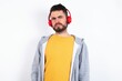 © Jihan - Serious displeased Young caucasian mán wearing trendy clothes over white background looks puzzled at camera being angry wears stereo headphones listens music while walking at street