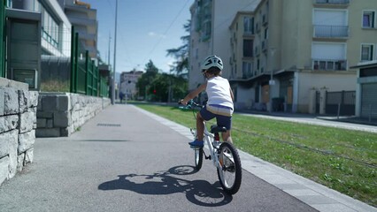 Wall Mural - Child rides bicycle in city street. Back of young boy cyclist riding bike outdoors in sidewalk urban environment