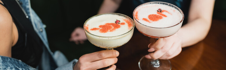 Wall Mural - Cropped view of multiethnic girlfriends holding cold foam cocktails in bar in evening, banner.