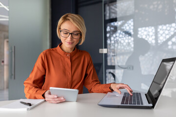 Young successful businesswoman at workplace is satisfied with results of achievement at work smiling and using tablet computer, blonde programmer is testing new software in application.