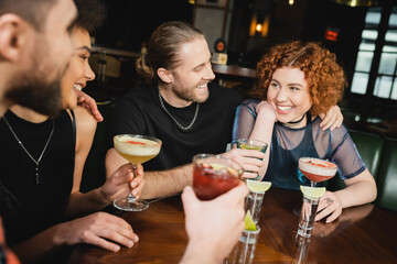 Wall Mural - Smiling multiethnic friends hugging and holding cocktails near tequila shots in bar.