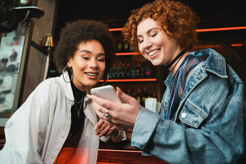 Wall Mural - Positive multiethnic friends using smartphone near stand in bar in evening.