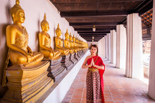 Young Woman Dressed In Traditional Red Thai Dress And Golden Ornaments Stands Sawasdee As A Welcome And Greeting In The Ancient Site With Many Buddha Row. Wat Phutthaisawan Ayutthaya