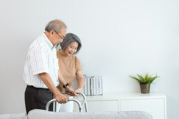 Wall Mural - Elderly health insurance, Asian woman support senior older man walk at home, help and take care of elderly mature patient to doing physical therapy with walker for body health in house living room