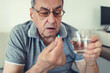 © Jelena Stanojkovic - Elderly man taking pills for depression sitting on couch. Old upset patient swallowing pill with glass of water.