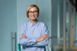 © Liubomir - Portrait of young successful business woman inside office at workplace, female worker standing with arms crossed smiling and looking at camera, woman in blue shirt and glasses blonde.