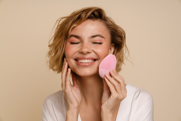 Cheerful woman using face cleansing brush isolated over beige background