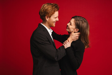 Beautiful couple hugging and looking at each other while standing isolated over red wall