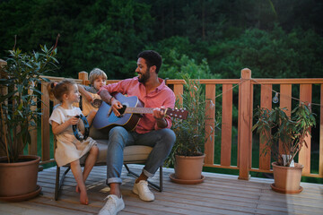 Young African american man enjoys playing the guitar with little children accompaining him with instruments on patio in garden in summer.