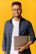 © Vadim Pastuh - Portrait of successful indian man in glasses standing and carrying laptop, looking at the camera isolated on yellow, cheerful male entrepreneur, freelancer or student smiling